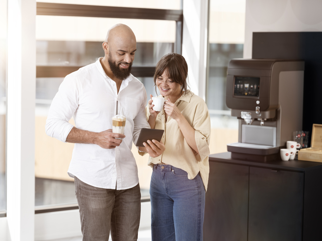 Deux personnes discutent près de la machine à café avec une tablette et des boissons dans un environnement de bureau lumineux.